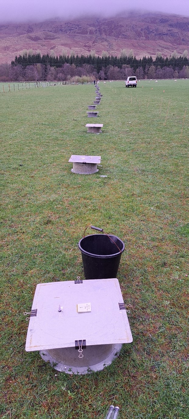 A series of grey, lidded chambers for nitrous oxide gas flux sampling arranged in a straight line across a grassy field. The closest chamber has binder clips securing its lid, and a black bucket with tubing sits nearby. A white van and a person are visible in the distance, with a backdrop of rolling hills and misty, tree-lined slopes